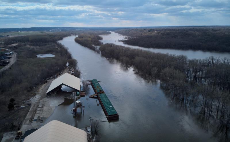 Aerial photo shows two channels of river with many trees in the water between them. A barge is docked near some large storage sheds that are partially underwater. 