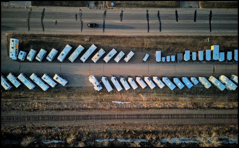 Looking straight down at a parking lot with two long rows of camper trailers for sale. There is a road above the lot and a railroad track is running underneath the lot.