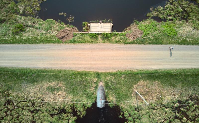 Looking straight down on a dirt road cutting the image in half horizontally. Bodies of water on either side of road are connected by a culvert. A tiny figure can be seen standing on the road casting a short shadow.