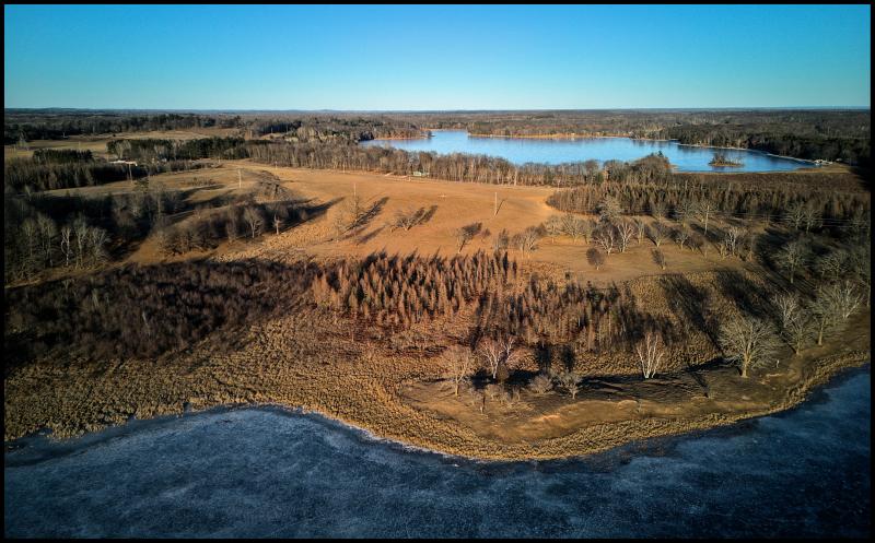 Aerial photo of some pastures between two frozen lakes. There is no snow on the ground and the fields are a soft brown, with patches of small trees casting shadows away from the camera.