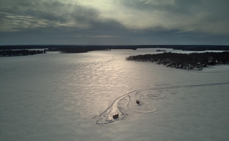 Drone photo from above of two tiny fish houses on a snow covered lake. It's a cloudy day, the sun is shining through and reflecting off the snow.