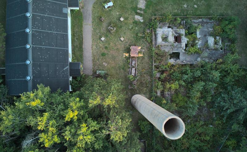 Looking straight down at an old mine site. We see the roof of a mid sized building on the left. The foundation of another building on the right and near the bottom center we are looking down a tall smoke stack. 