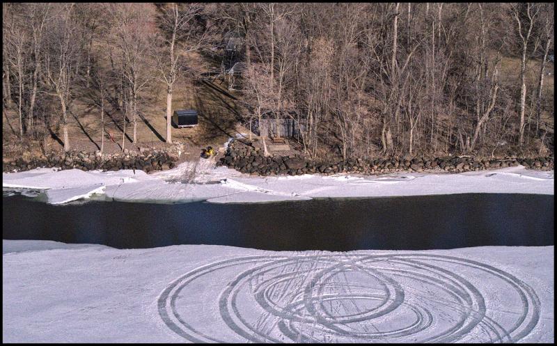Aerial view of shoreline with some snow on shore, some open water, and then ice. On the ice one can see snow mobile tracks spinning in a circle. The path to shore is interrupted by open water and a shift by the ice to the left.