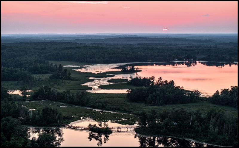 Aerial view overlooking two neighboring lakes with swamps between them. It's sunsent and the colors are reflecting across one lake, in the other we see the reflection of a walkway across the lake. 