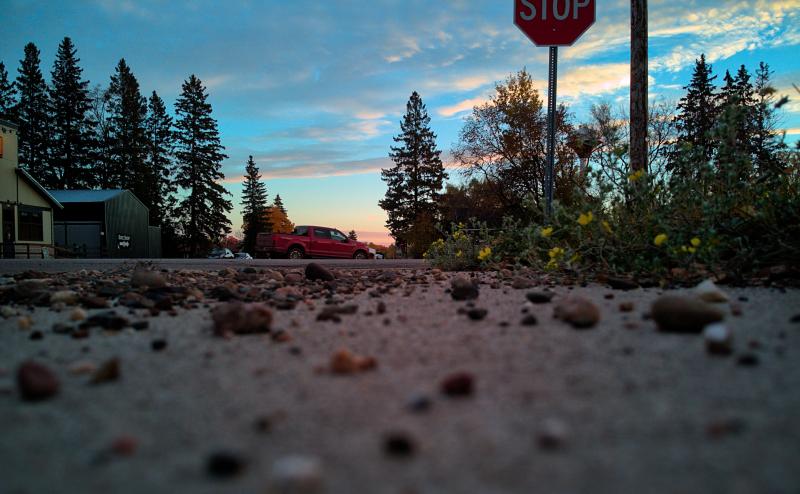Photo taken from about one inch above the pavement shows many small pebbles that look quite large, with a stop sign above and a red pickup truck in the distance. 