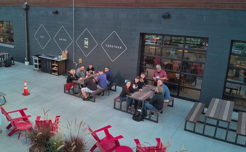 Two tables on a patio with folks socializing, drinking beer, and a few are waving to the camera.