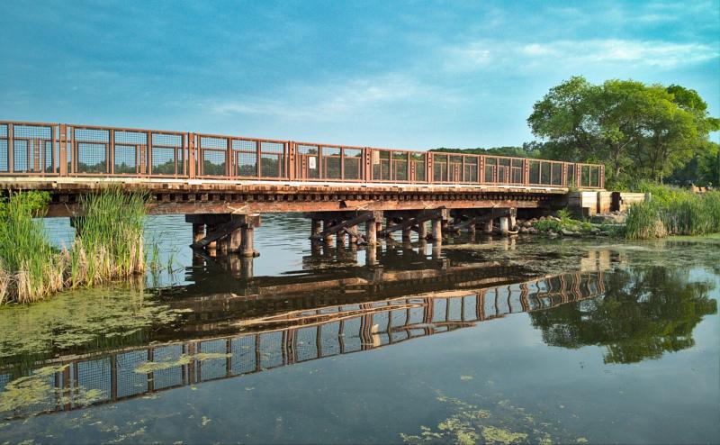 An low bicycle bridge on wooden legs passes over a body of water. The sky is very blue and the brige is reflecting in the water. 