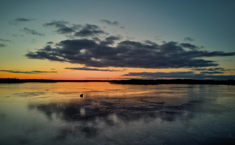 Photo of sunset over a frozen lake with the small silouette of a very small ice house and 4 wheeler in the middle of the frozen lake.
