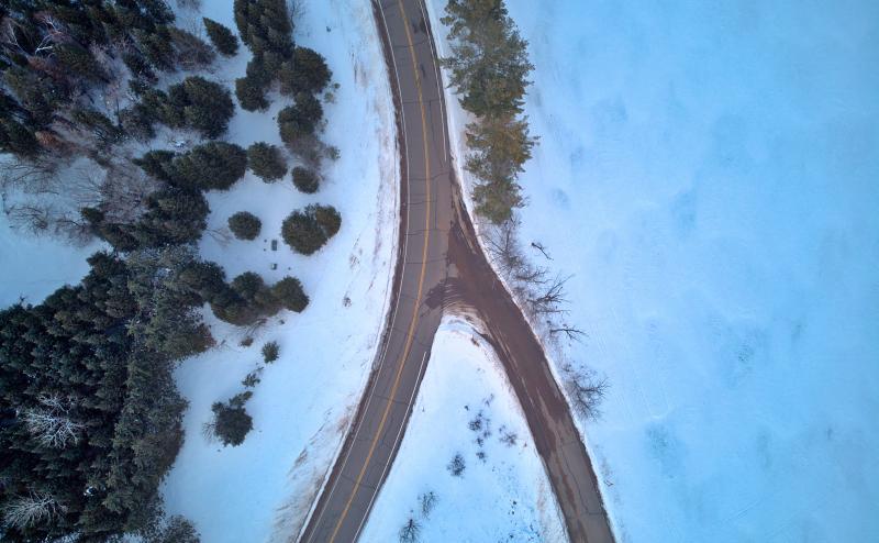 Looking straight down on a bare road where it comes to a Y in a snowcovered landscape. Trees on left side of image and what appears to be a frozen and snow covered lake on the right side.